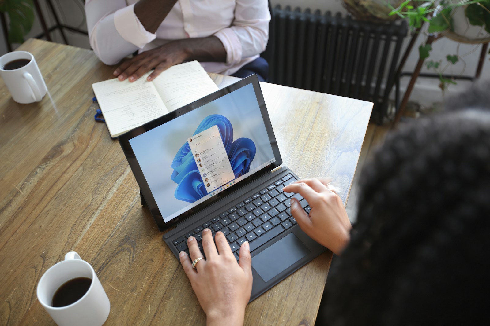 Overhead view of two people at a table working with a Microsoft laptop and notebook — Acronis Bulut Yedekleme: İş verilerinizi %100 güvende tutun.
